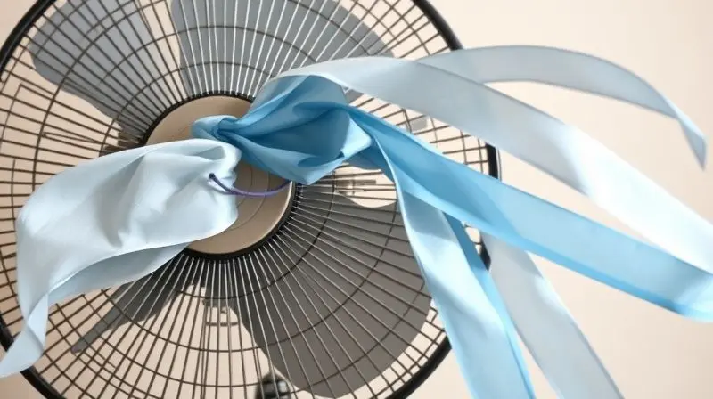 Fitas de seda azul e branca presas à grade de um ventilador, esticadas horizontalmente pela força do vento, ilustrando a potência do aparelho.